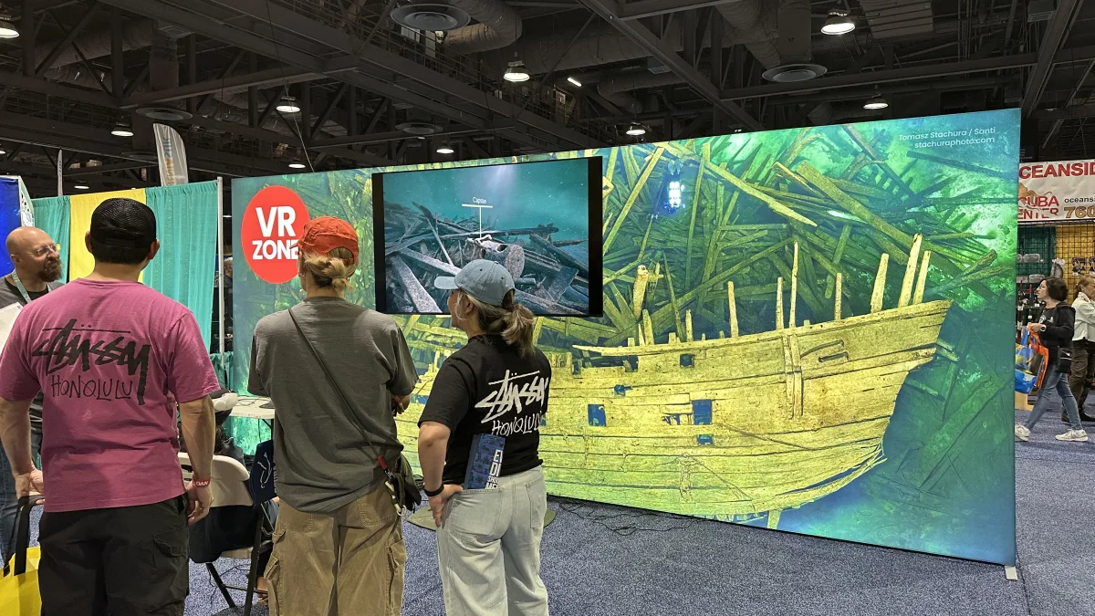 Indoor expo/trade-show booth scene with a large illustrated underwater shipwreck backdrop and a monitor displaying an underwater image; several attendees stand in the foreground viewing the display.