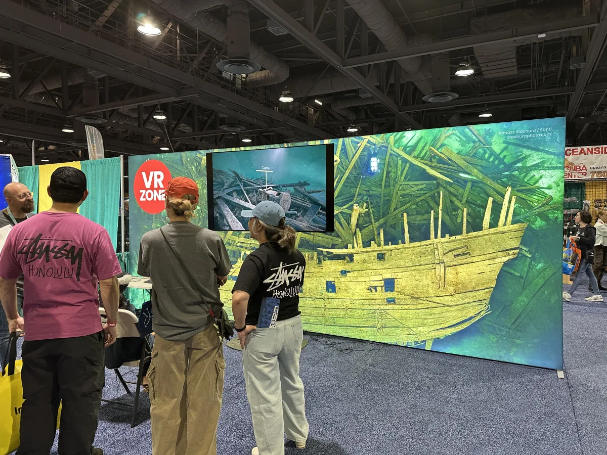 Indoor expo/trade-show booth scene with a large illustrated underwater shipwreck backdrop and a monitor displaying an underwater image; several attendees stand in the foreground viewing the display.