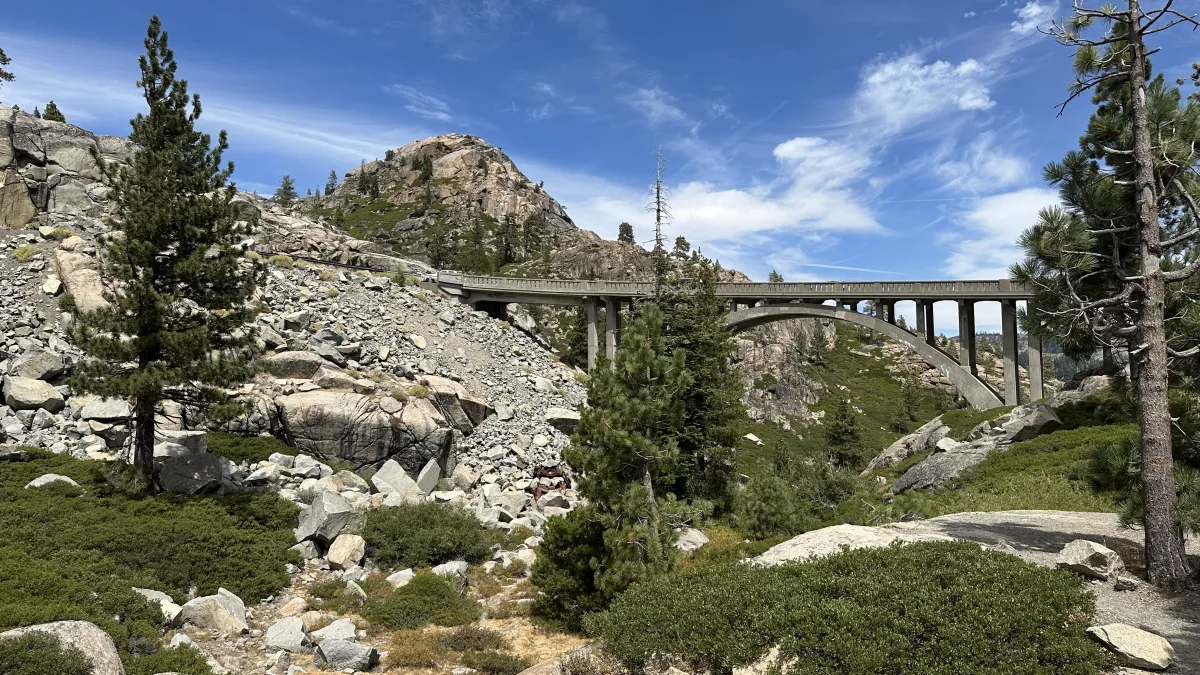 Wide landscape photo of a rocky mountainous area with scattered evergreen trees and a concrete arch bridge spanning a ravine under a bright blue sky with thin clouds.