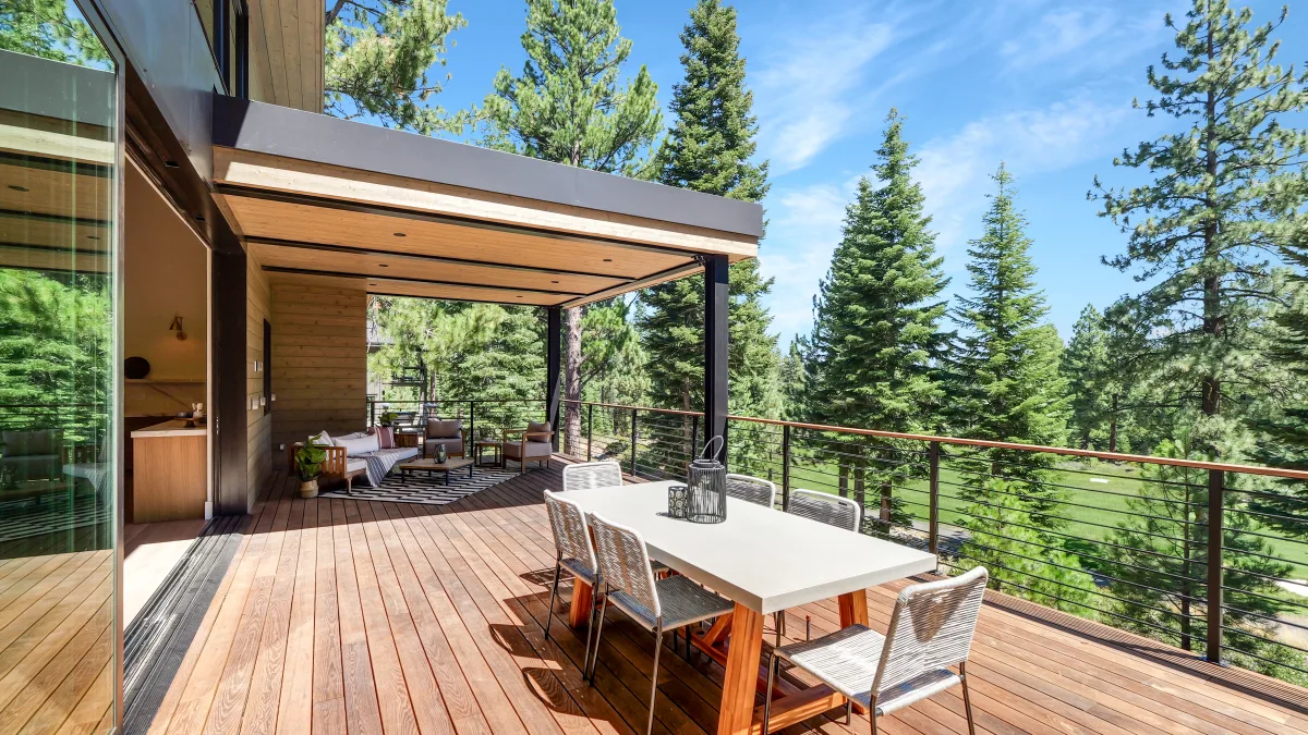 Modern outdoor deck/terrace attached to a contemporary home, featuring a dining table with chairs and a lounge seating area under an overhang, overlooking a forested landscape of tall evergreen trees under a clear blue sky.