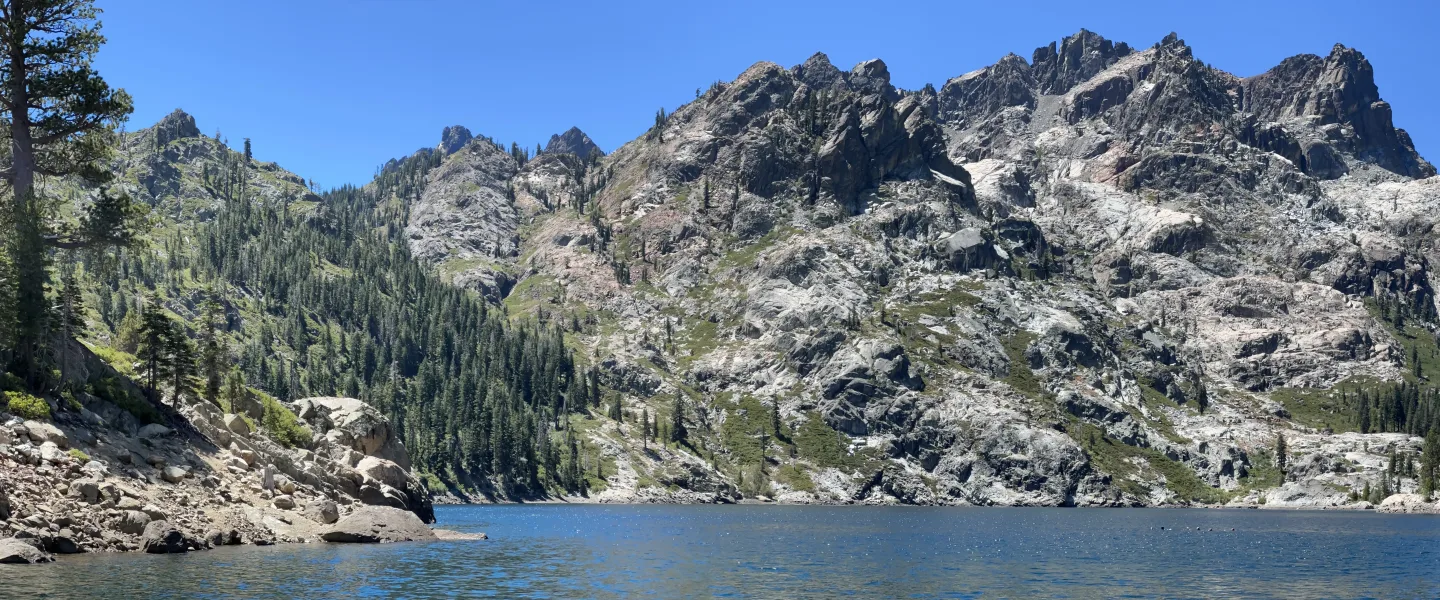 Wide panoramic landscape photo of a mountain lake with clear blue water in the foreground, rocky granite peaks and forested slopes in the background, and a cloudless blue sky.