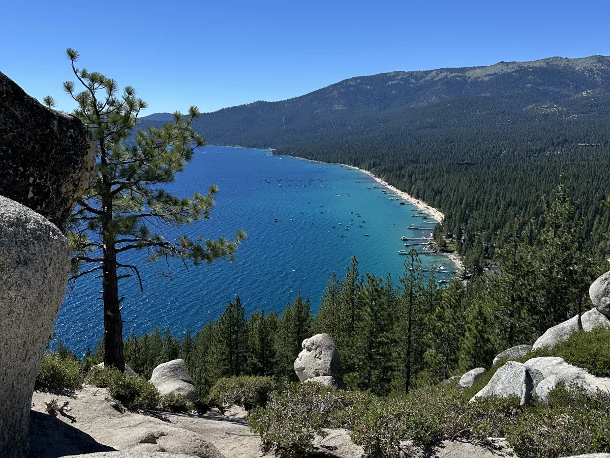 Panoramic lakeside landscape viewed from a rocky overlook: a deep-blue lake curves along a forested shoreline with a sandy beach, small docks, and many boats; pine trees and boulders frame the foreground under a clear blue sky.