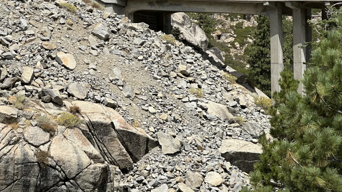 A concrete bridge/overpass spans a rocky mountain slope with granite boulders and scattered evergreen trees under a bright sky.