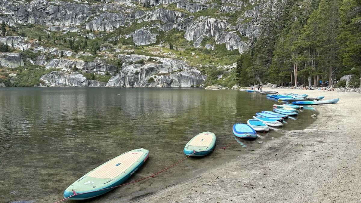 Lakeside scene with a sandy beach in the foreground, multiple stand-up paddleboards lined up and tethered along the water’s edge, and a backdrop of rocky granite slopes with scattered evergreen trees.