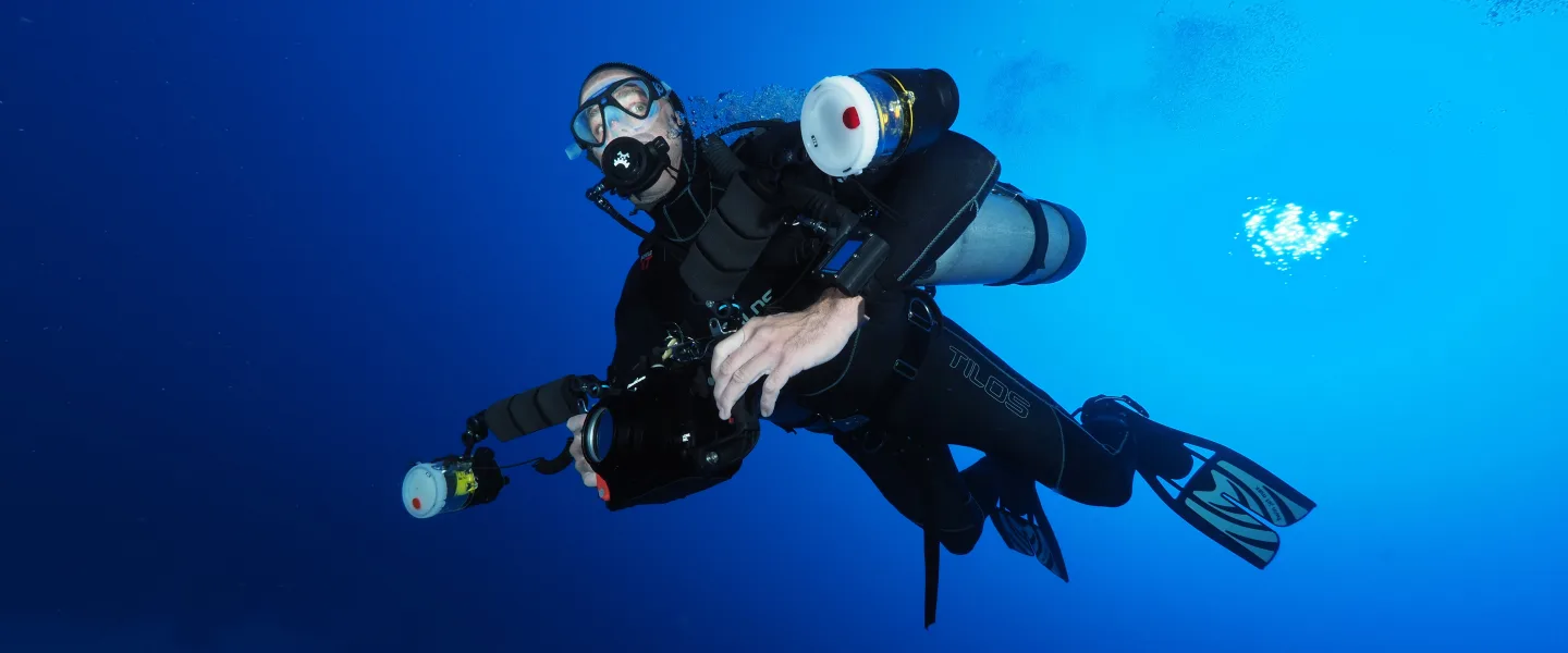 Underwater wide shot of a scuba diver in open blue water, wearing a mask and regulator and carrying multiple cylinders and underwater camera/lighting equipment; bubbles and light reflections are visible in the water.