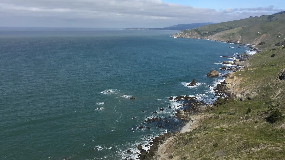 Wide coastal landscape photo showing an ocean shoreline with rocky cliffs and green hills on the right, open water to the left, and a cloudy band across a blue sky.