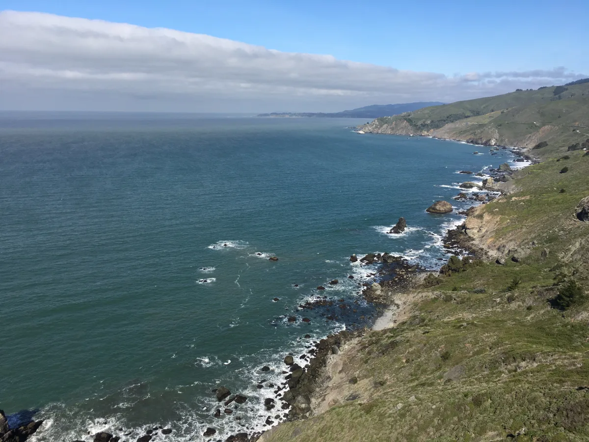 Wide coastal landscape photo showing an ocean shoreline with rocky cliffs and green hills on the right, open water to the left, and a cloudy band across a blue sky.