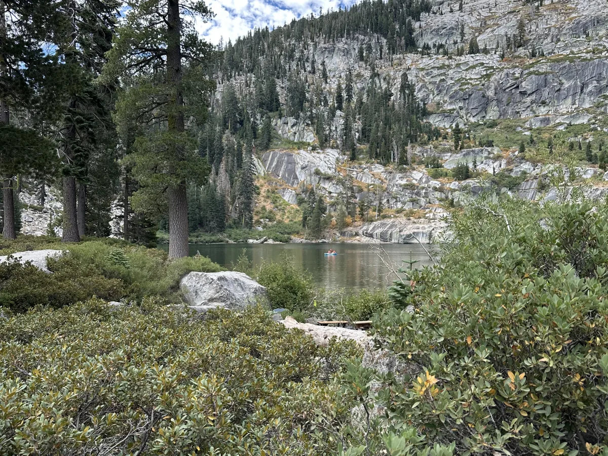Wide outdoor landscape showing a forested rocky mountainside beyond a calm lake, with shrubs and boulders in the foreground and a small boat on the water under a partly cloudy blue sky.