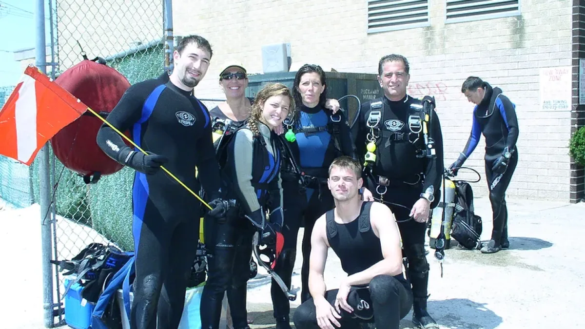 Group photo of scuba divers in wetsuits and full dive gear posing outdoors near a chain-link fence and a beige brick building; a diver holds a red-and-white diver-down flag on a pole, with bags and equipment on the ground and one person in