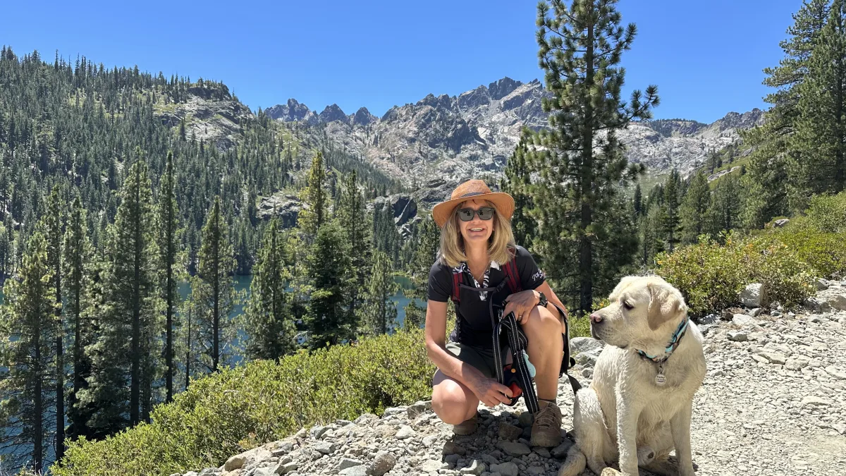 Outdoor portrait-style photo of a hiker kneeling on a rocky trail next to a light-colored dog, with a pine-forested mountain landscape, rocky peaks, and a lake visible in the background under a clear blue sky.