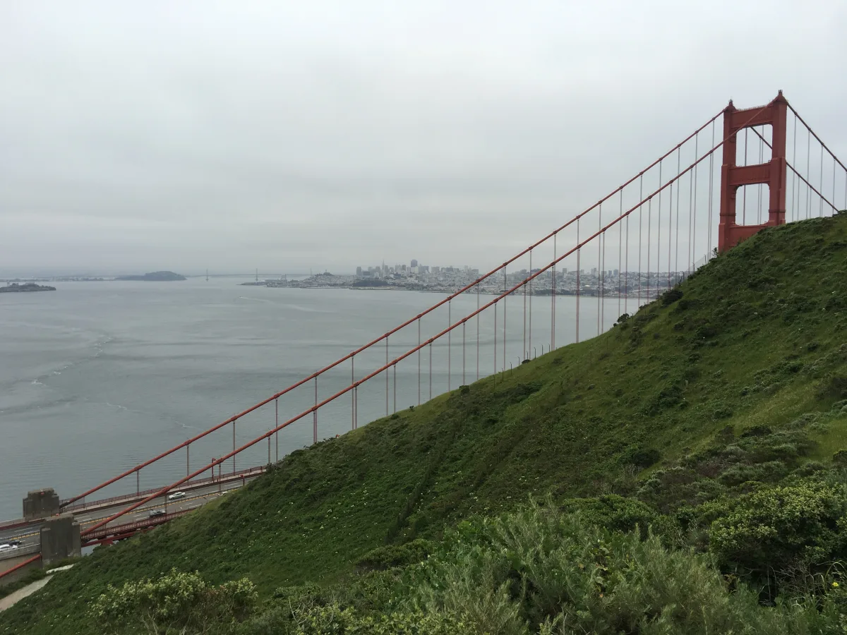 Wide landscape photo showing a large red suspension bridge tower and cables on the right, spanning over a bay with a distant city skyline under an overcast sky.