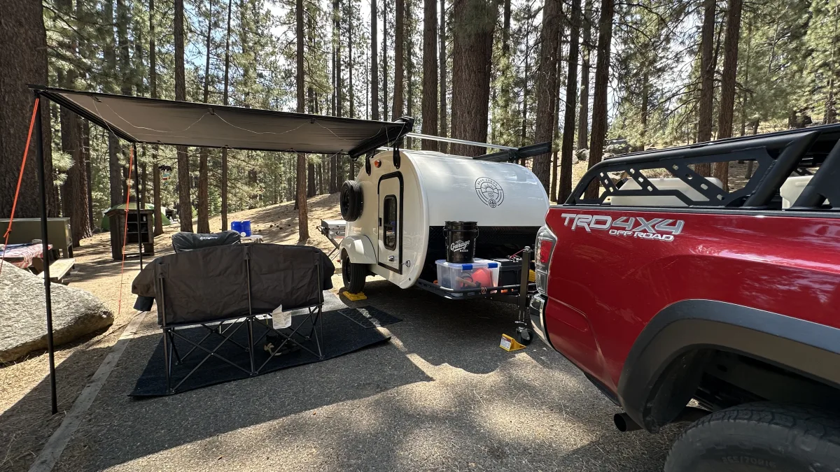 Outdoor campsite scene in a pine forest with a red pickup truck hitched to a small white teardrop-style travel trailer.