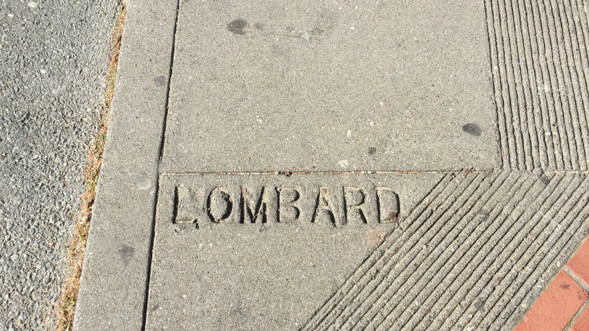 Close-up, downward-facing photo of a concrete sidewalk corner/curb ramp area with the word "LOMBARD" engraved in the concrete near the bottom; adjacent surfaces include asphalt on the left and a grooved concrete ramp with red brick edging o