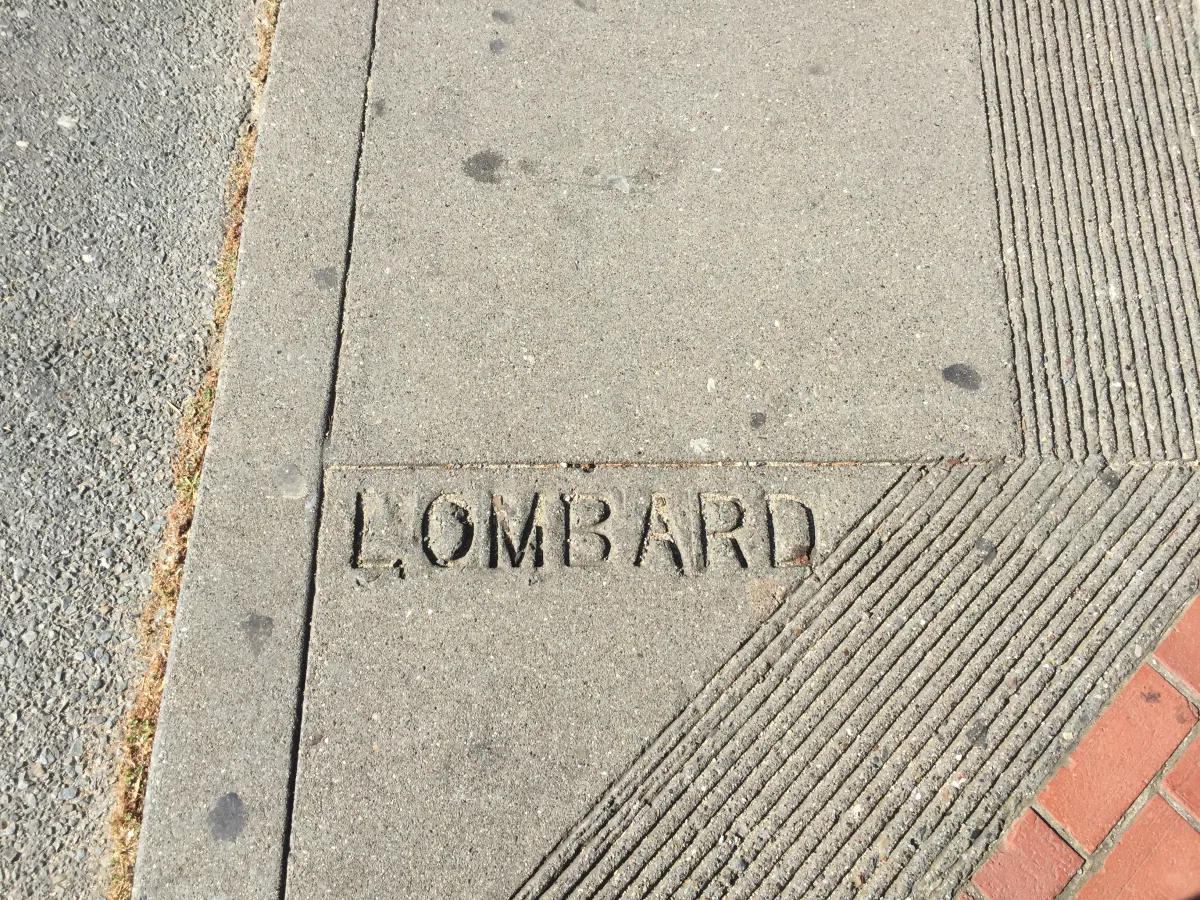 Close-up, downward-facing photo of a concrete sidewalk corner/curb ramp area with the word "LOMBARD" engraved in the concrete near the bottom; adjacent surfaces include asphalt on the left and a grooved concrete ramp with red brick edging o