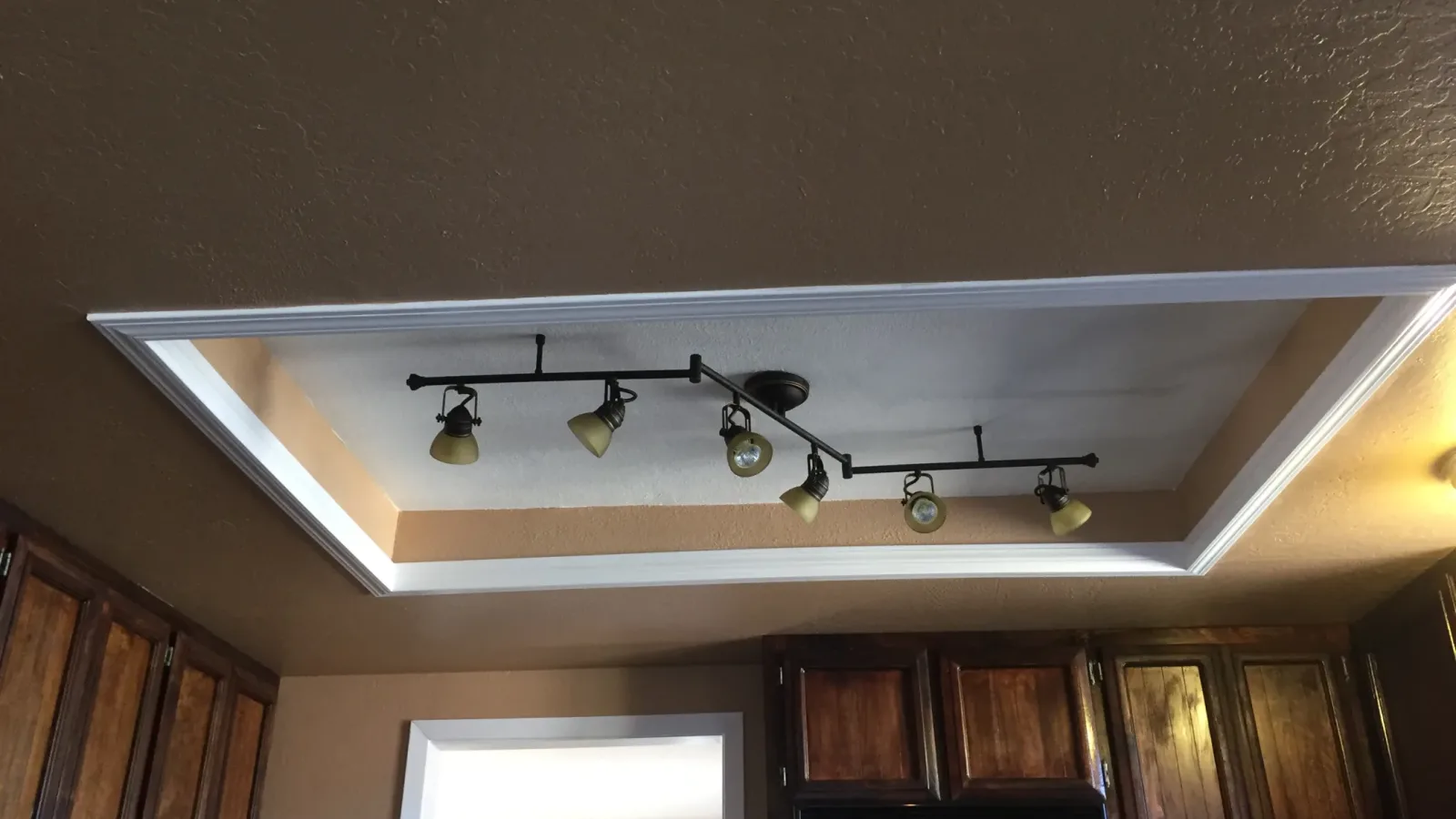 Interior photo looking upward at a recessed tray ceiling detail with white crown molding and a black multi-head track light fixture; upper portions of dark wood kitchen cabinets and part of a stainless microwave are visible at the bottom ed