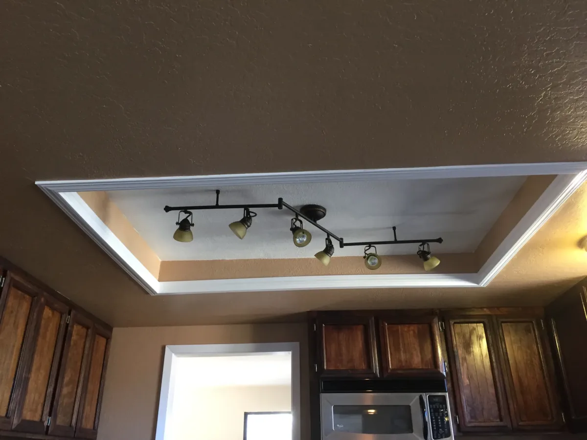 Interior photo looking upward at a recessed tray ceiling detail with white crown molding and a black multi-head track light fixture; upper portions of dark wood kitchen cabinets and part of a stainless microwave are visible at the bottom ed