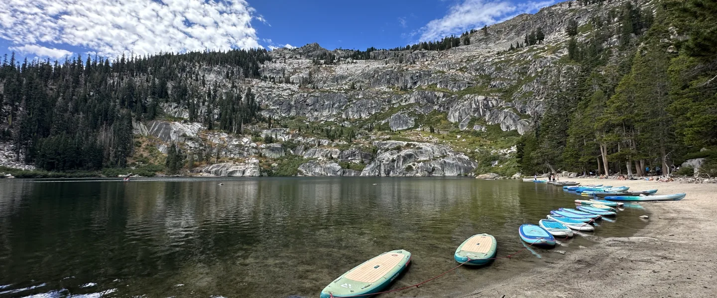 Wide landscape photo of a calm mountain lake with a sandy shoreline in the foreground.