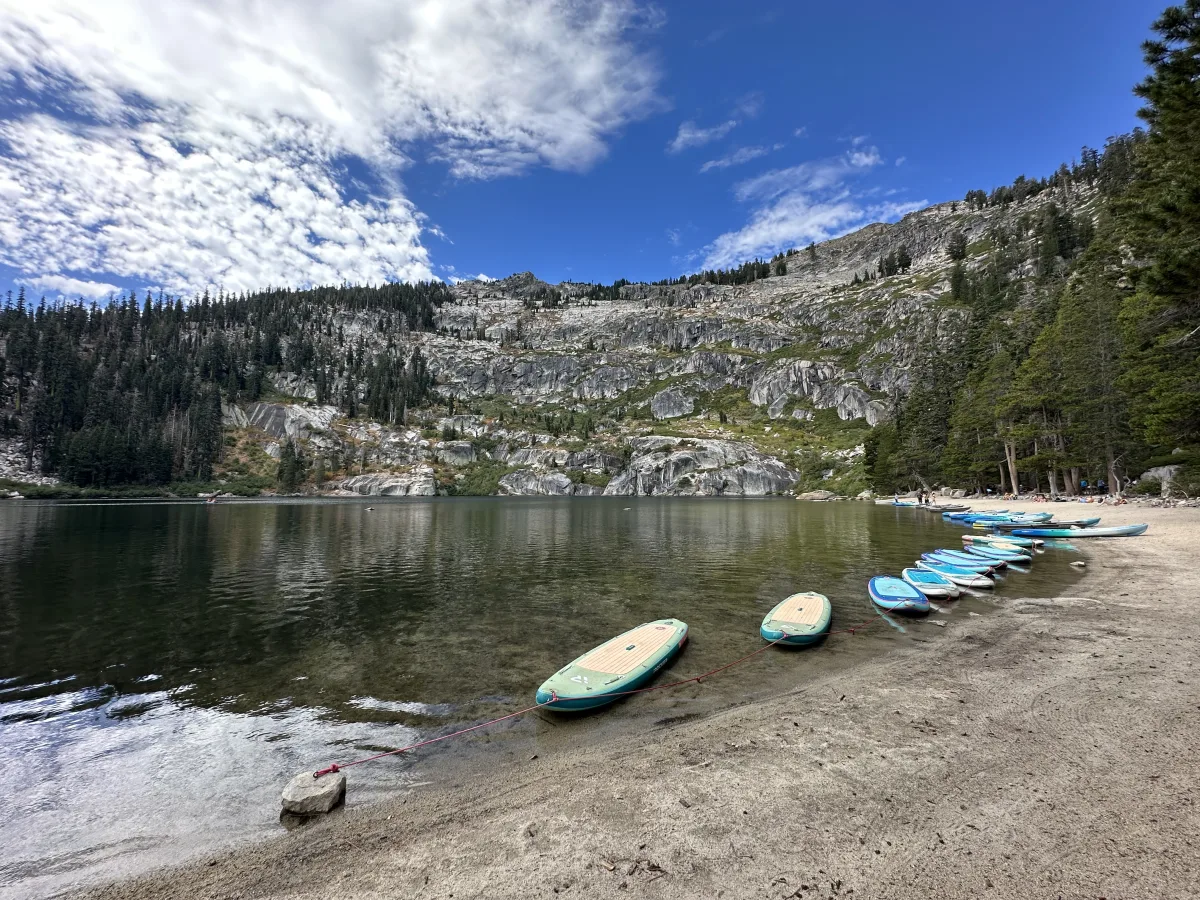 Wide landscape photo of a calm mountain lake with a sandy shoreline in the foreground.