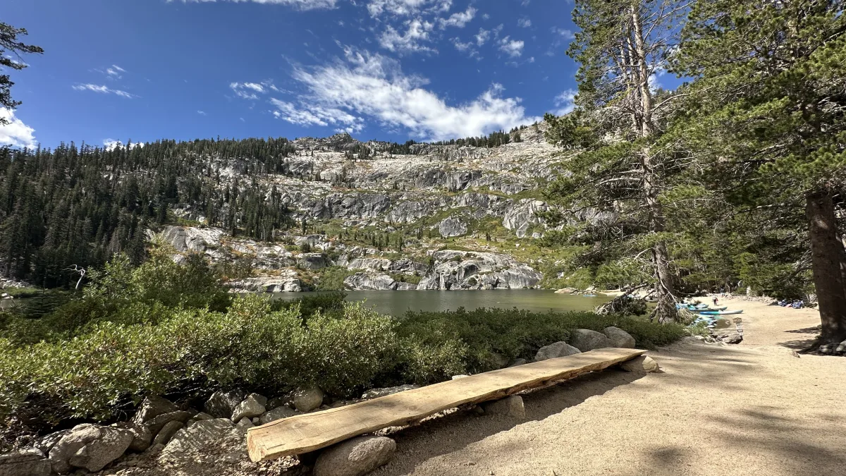 Wide outdoor landscape photo showing a mountain lake with rocky granite slopes and dense conifer forest under a blue sky with scattered clouds.