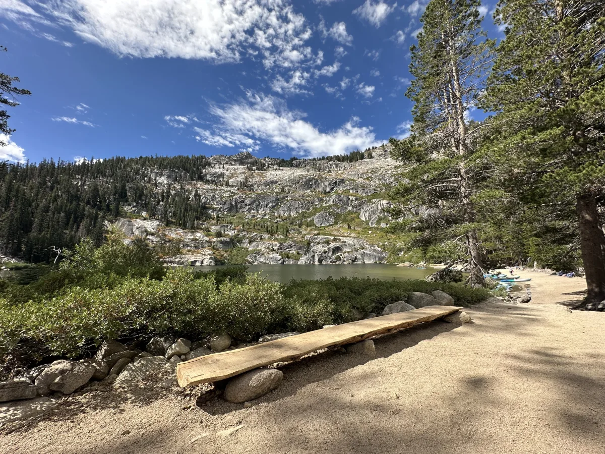 Wide outdoor landscape photo showing a mountain lake with rocky granite slopes and dense conifer forest under a blue sky with scattered clouds.