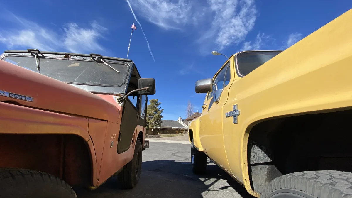 Low-angle outdoor photo looking up between two parked vintage vehicles: an older orange Jeep on the left and a yellow Chevrolet pickup truck on the right, with a deep blue sky and wispy clouds overhead; a small American flag is visible abov