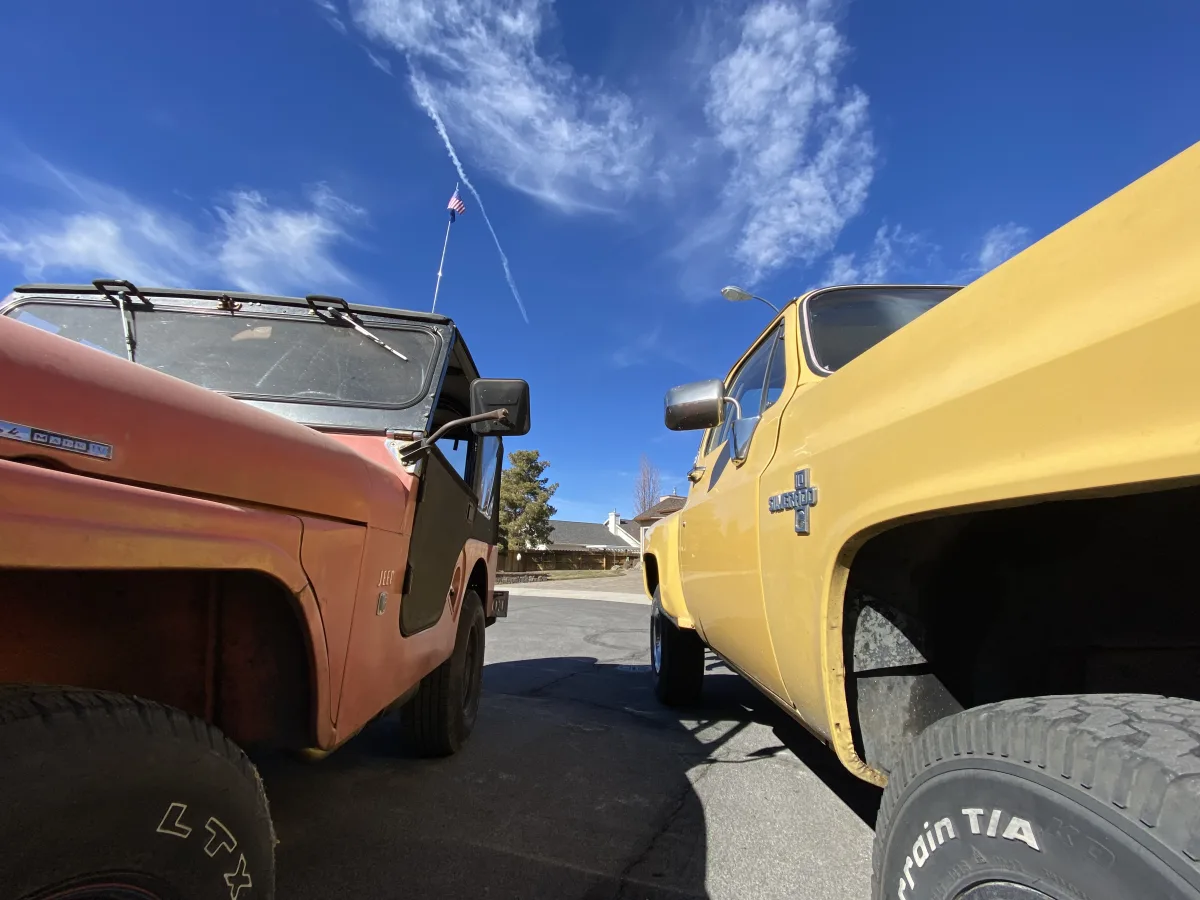 Low-angle outdoor photo looking up between two parked vintage vehicles: an older orange Jeep on the left and a yellow Chevrolet pickup truck on the right, with a deep blue sky and wispy clouds overhead; a small American flag is visible abov