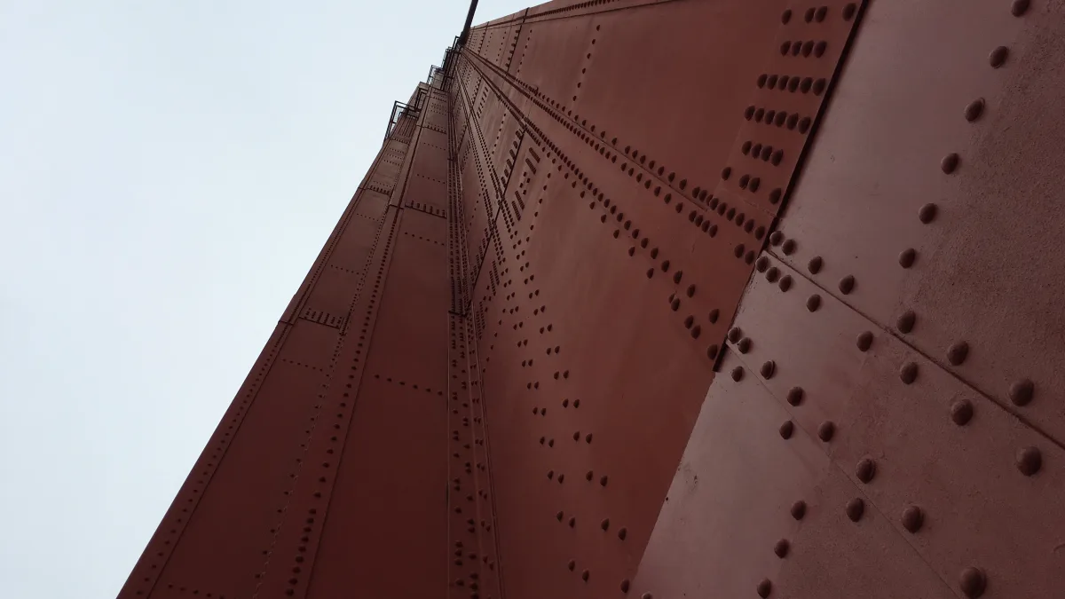 Low-angle close-up view of a large riveted metal structure painted reddish-brown, with repeating rows of round rivets and vertical/diagonal steel members; pale sky fills the left background.