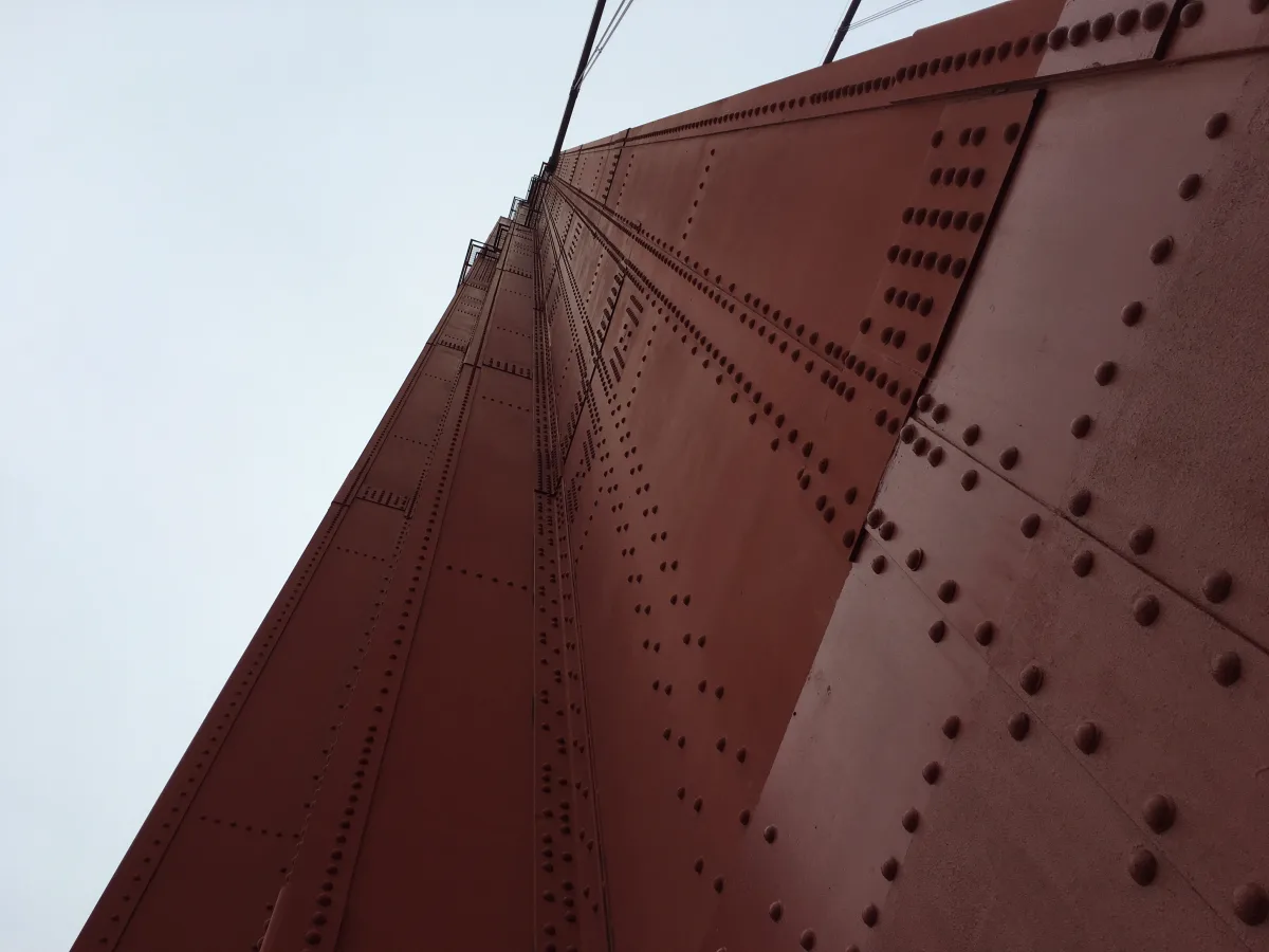 Low-angle close-up view of a large riveted metal structure painted reddish-brown, with repeating rows of round rivets and vertical/diagonal steel members; pale sky fills the left background.