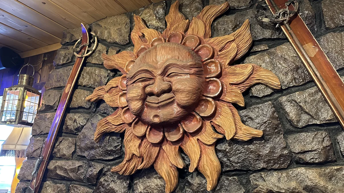 Rustic interior wall featuring a large carved wooden sun face mounted on dark stone masonry, flanked by two vintage wooden skis; a metal-and-glass lantern light fixture appears on the left under a wood plank ceiling.
