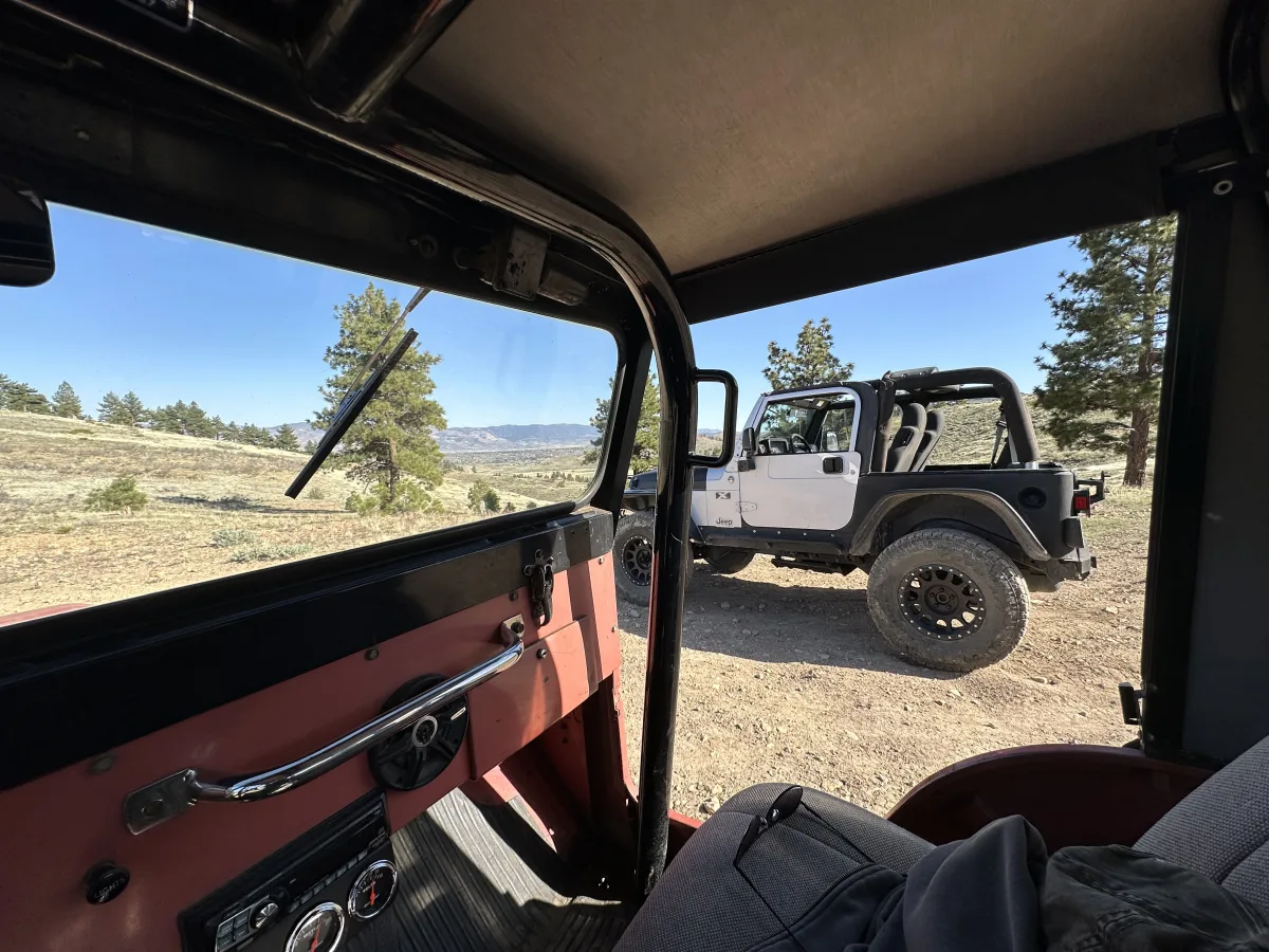 View from inside a vehicle looking out through an open side toward a white off-road Jeep-style SUV parked on a dirt pull-off in a hilly landscape with pine trees and distant mountains under a clear blue sky.