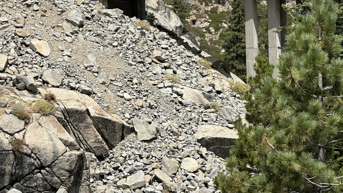 Outdoor mountain scene featuring a concrete road bridge spanning a rocky slope/ravine, with granite boulders, sparse shrubs, and pine trees under a lightly cloudy sky.