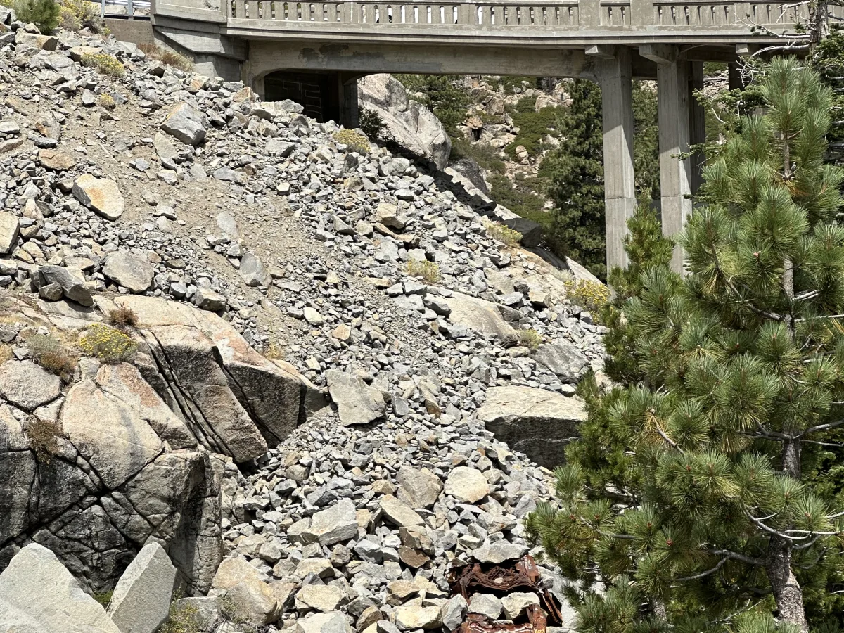 Outdoor mountain scene featuring a concrete road bridge spanning a rocky slope/ravine, with granite boulders, sparse shrubs, and pine trees under a lightly cloudy sky.