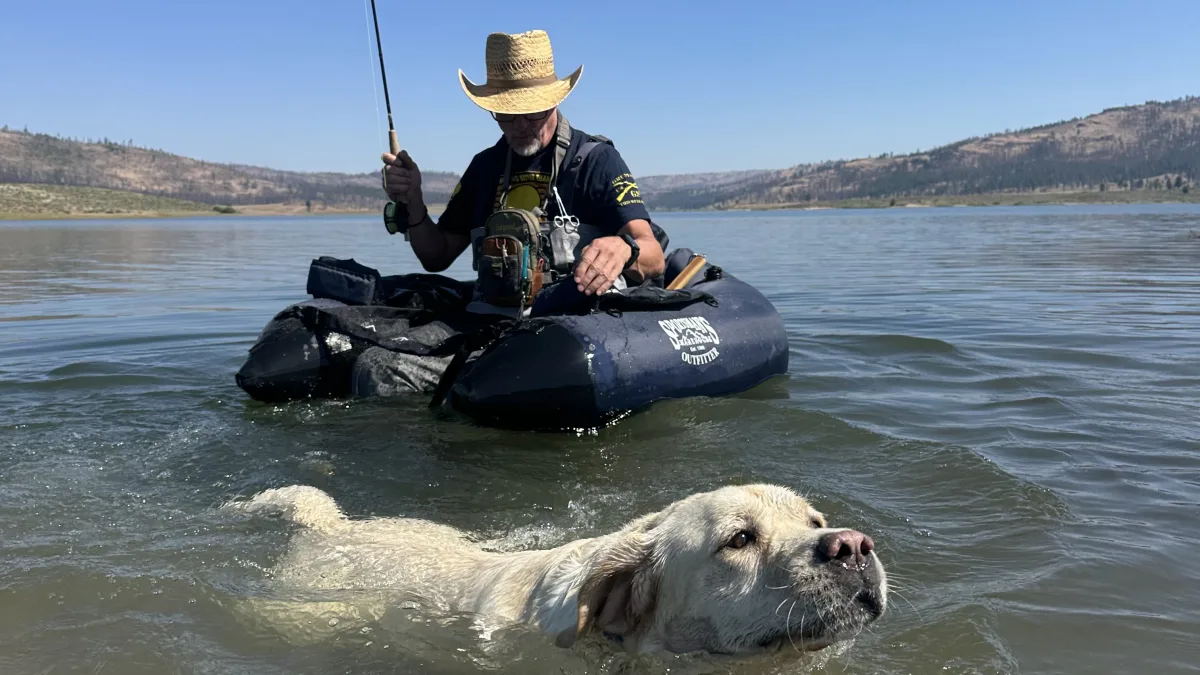 Outdoor lake scene with a person seated in a dark inflatable float tube holding a fishing rod, while a light-colored dog swims in the foreground.