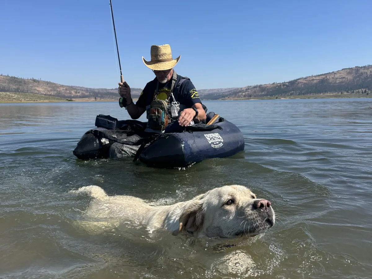 Outdoor lake scene with a person seated in a dark inflatable float tube holding a fishing rod, while a light-colored dog swims in the foreground.