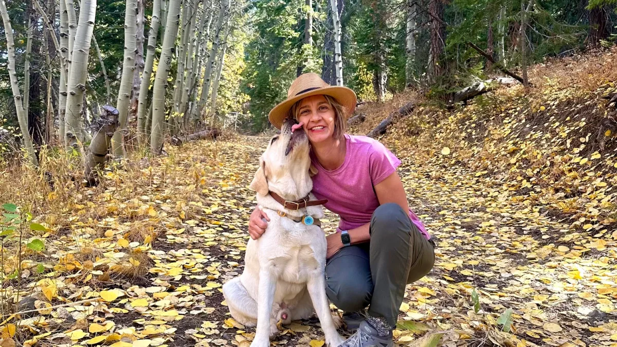 Outdoor portrait-style photo of a person crouching on a leaf-covered trail in a wooded area, hugging a light-colored dog that is licking the person’s face.
