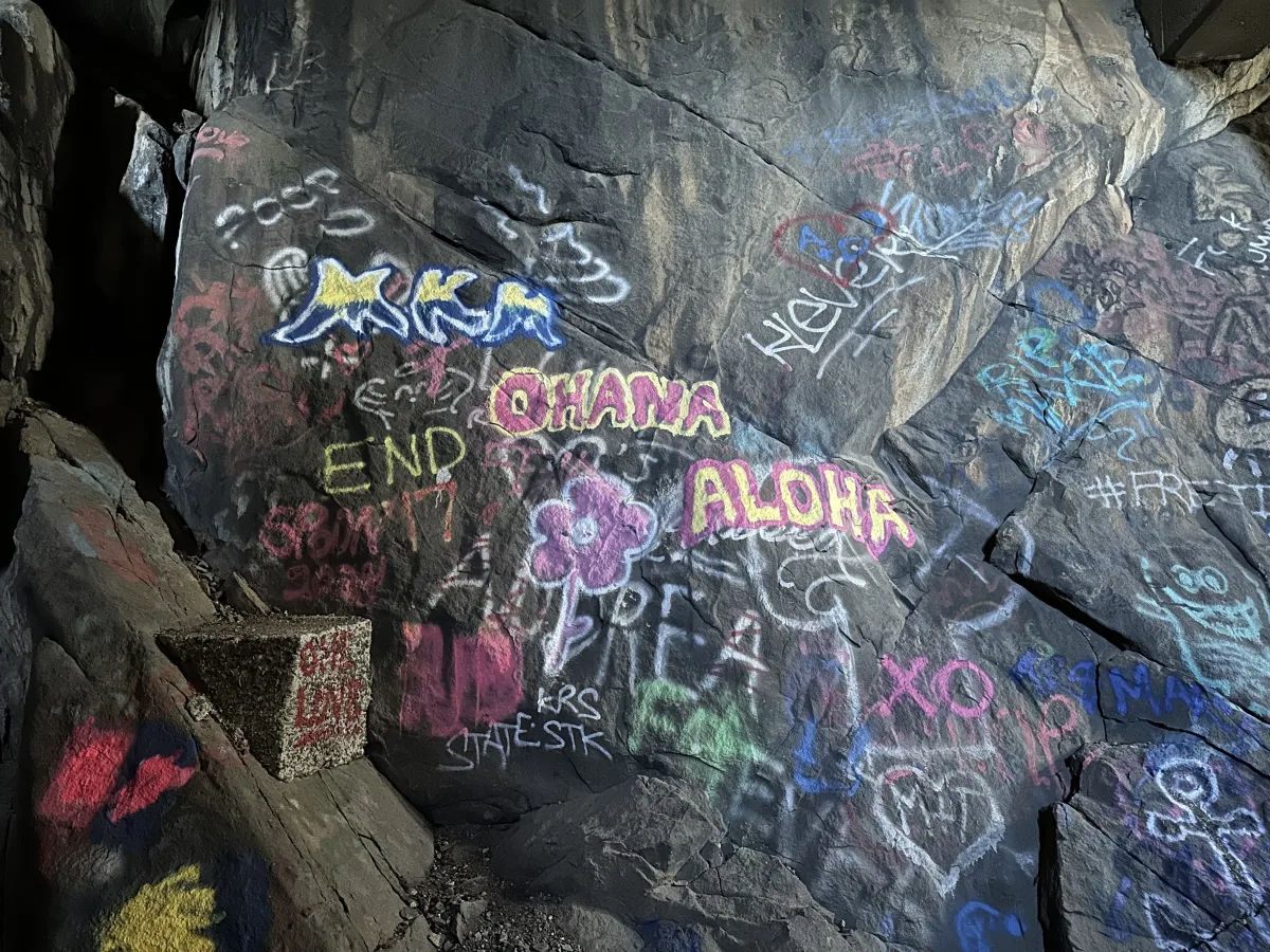 A large dark rock wall covered with colorful spray-painted graffiti, including stylized words and a flower drawing, in what appears to be a rocky, cave-like or underpass setting.