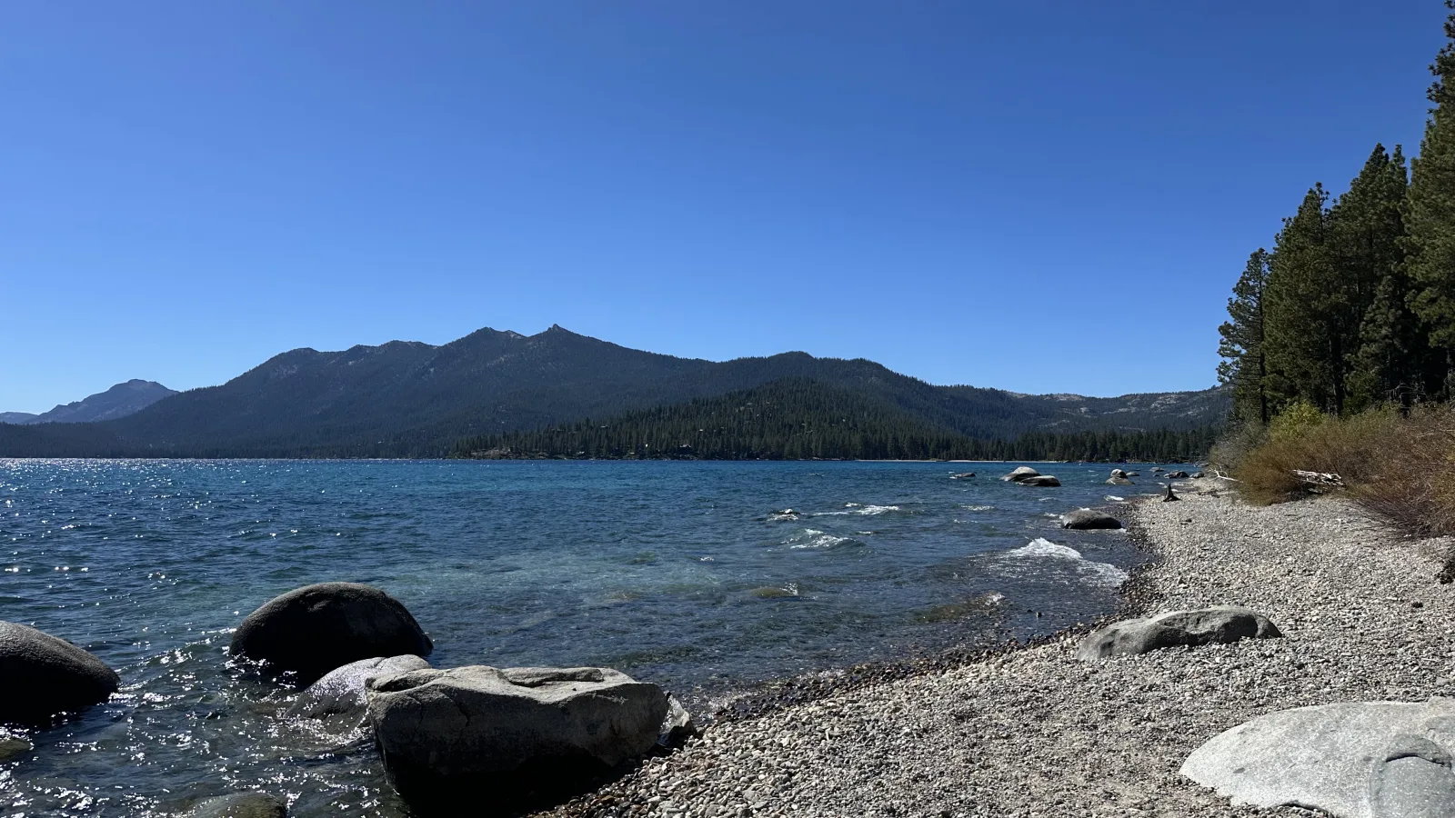 Wide landscape photo of a calm lakeshore with clear blue sky, a forested mountain ridge in the distance, and a rocky/pebble beach in the foreground with large boulders near the waterline.