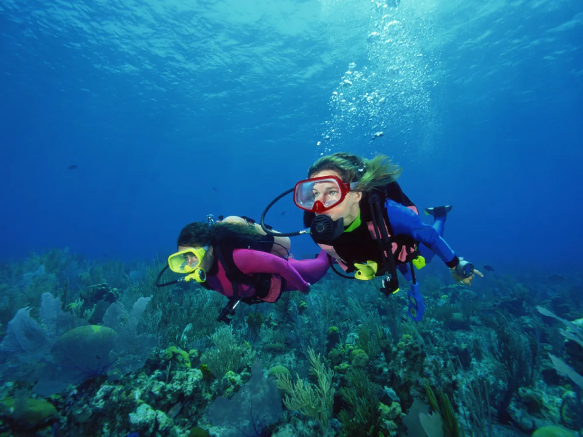 Underwater photo of two scuba divers swimming above a coral reef, with open blue water and bubbles rising toward the surface.