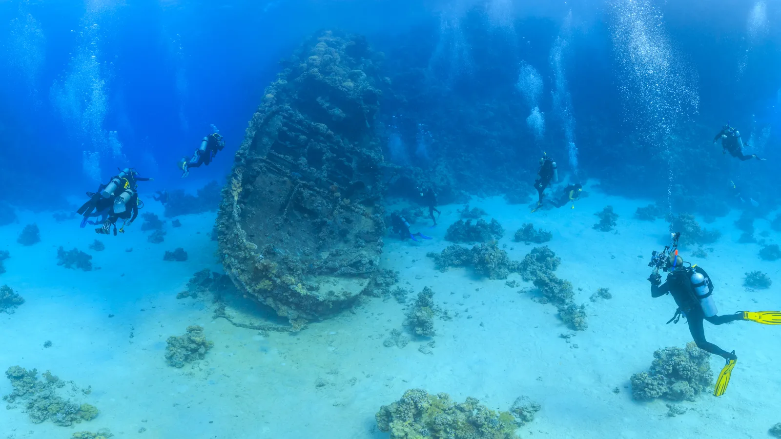 Wide underwater scene showing multiple scuba divers exploring a large, coral-encrusted shipwreck resting on a sandy seabed, with air bubbles rising through blue water.