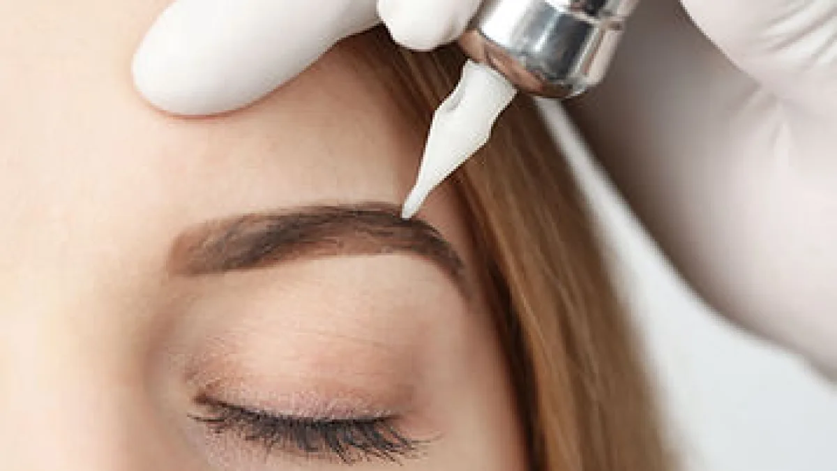 Close-up of a person’s closed eye and eyebrow while a gloved hand uses a pen-like cosmetic tool near the eyebrow, suggesting a brow/skin treatment being performed.