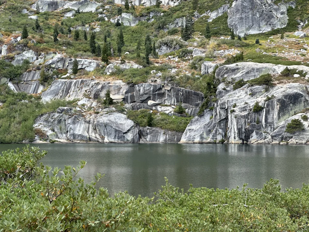 Landscape photo showing a calm body of water in the foreground and a rocky hillside in the background with large gray granite outcrops, scattered conifer trees, and low shrubs.