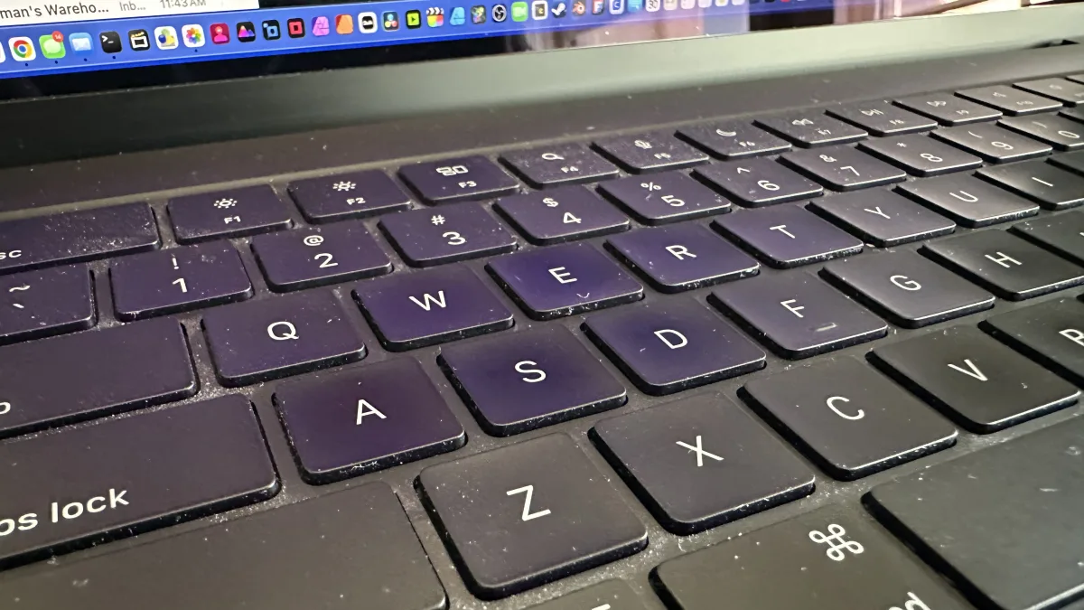 Close-up, angled photo of a laptop keyboard in the foreground with part of the laptop screen visible at the top showing a macOS-style dock and a browser/web app interface.