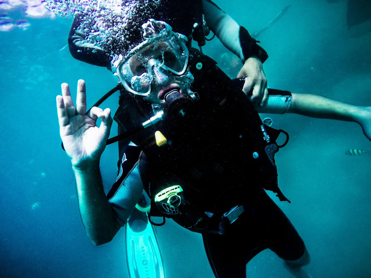 Underwater close-up photo of a scuba diver in full gear, facing the camera and making an “OK” hand signal, with bubbles rising and a second diver partially visible behind.
