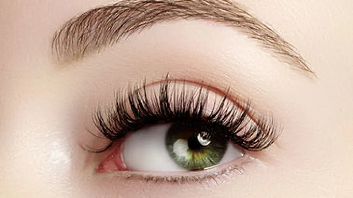 Close-up beauty photo showing a single human eye with long, styled eyelashes and a well-defined eyebrow against smooth, light-toned skin.