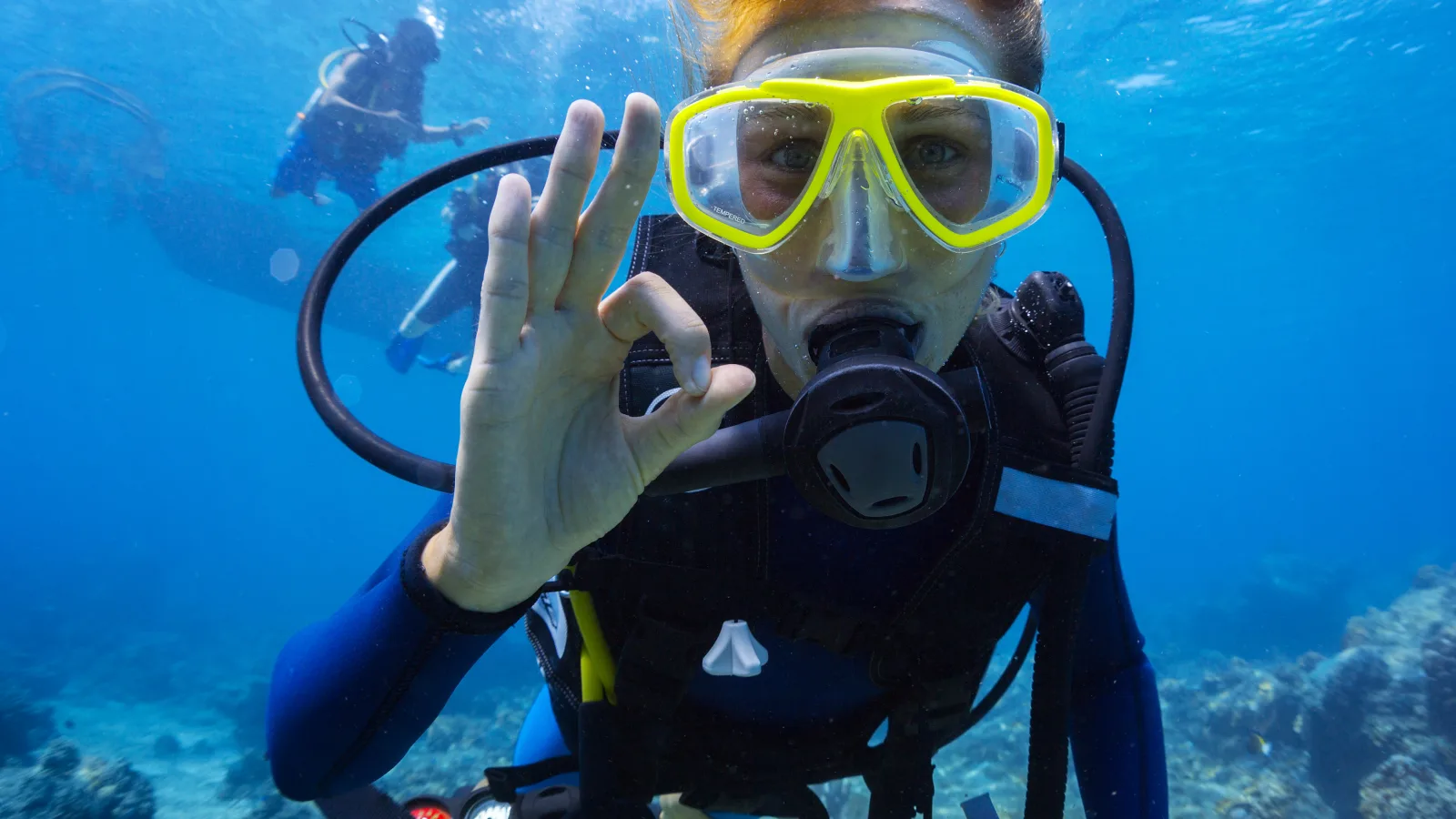 Underwater close-up of a scuba diver wearing a yellow mask and breathing regulator, giving an “OK” hand signal; another diver appears blurred in the background over a blue ocean scene with bubbles and a rocky seabed.