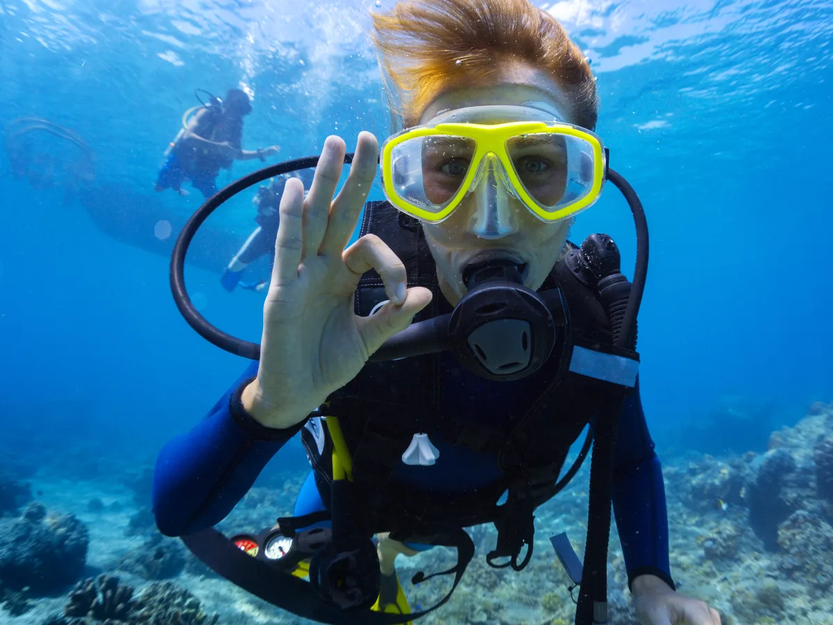 Underwater close-up of a scuba diver wearing a yellow mask and breathing regulator, giving an “OK” hand signal; another diver appears blurred in the background over a blue ocean scene with bubbles and a rocky seabed.