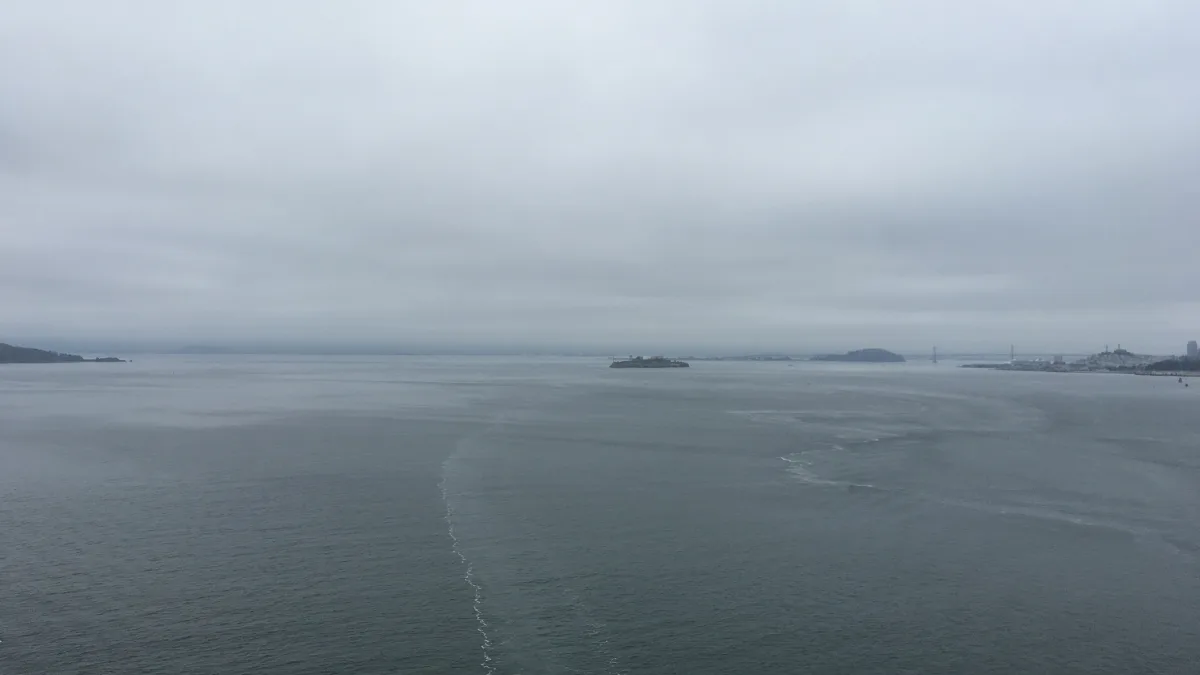 Wide, overcast seascape with calm water, a faint wake line leading toward the horizon, and distant landforms including a small island and a far shoreline with a bridge-like structure on the right.