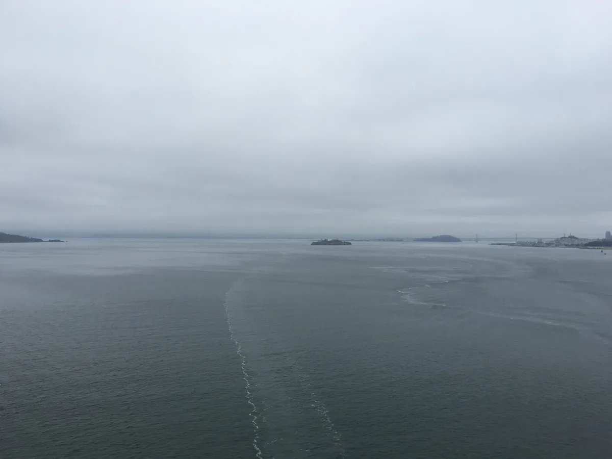Wide, overcast seascape with calm water, a faint wake line leading toward the horizon, and distant landforms including a small island and a far shoreline with a bridge-like structure on the right.