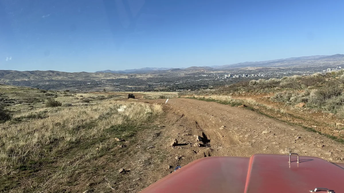 Wide landscape view from a hillside dirt road/trail with scrubby grass and rocks, overlooking a valley and a distant city skyline backed by mountain ridges under a clear blue sky.