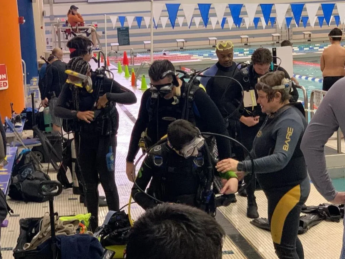 Indoor swimming pool scene with a group of people in scuba gear (wetsuits, BCDs, tanks, masks) preparing on the pool deck; lanes and blue triangular pennant flags are visible in the background.